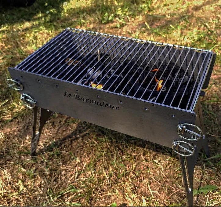 Barbecue portable en métal avec grille de cuisson "Le Baroudeur", posé sur de l'herbe, utilisé pour faire des grillades.