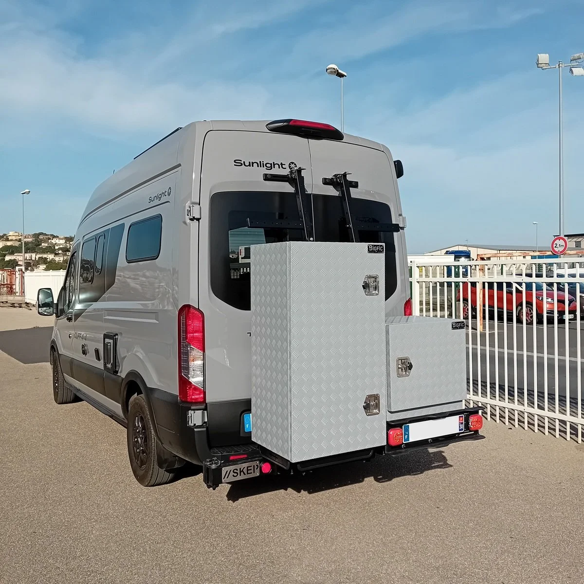 Un véhicule utilitaire blanc équipé de coffres en aluminium BIGFIC, stationné dans un parking, avec un environnement urbain en arrière-plan.
