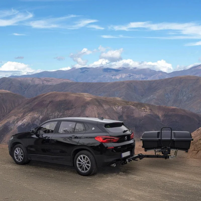 Une voiture noire avec un coffre de remorque stationnée sur une route de montagne, avec des montagnes et un ciel bleu en arrière-plan.