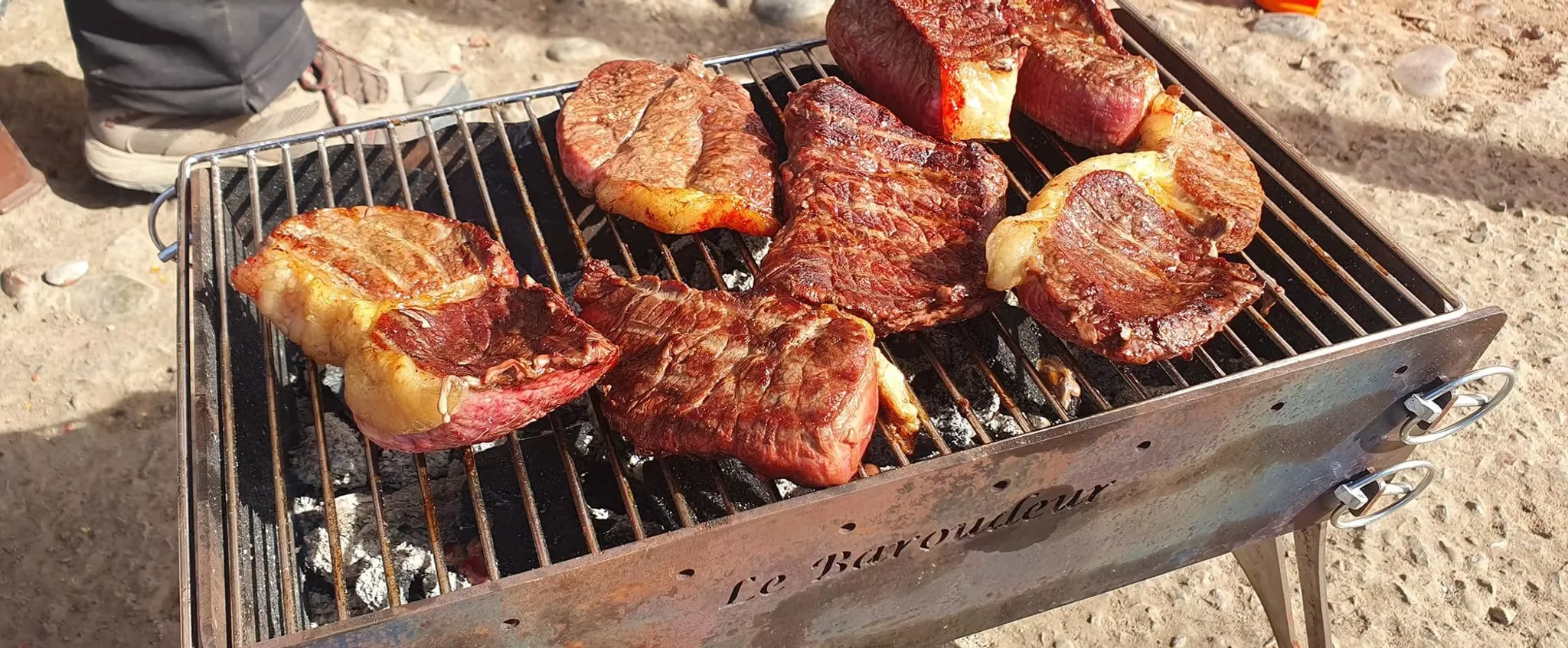 Des morceaux de viande en train de griller sur un barbecue en extérieur sur un sol sableux.