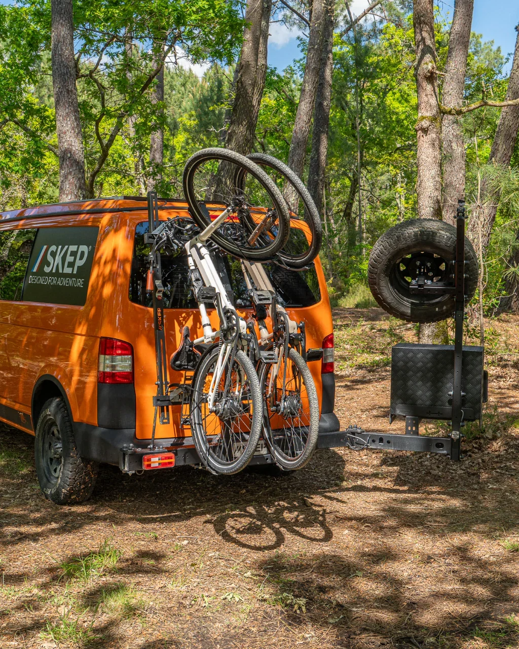 Un van orange équipé avec un porte-matériel Skep pour le transport de vélos, stationné dans une forêt, avec des vélos fixés à l'arrière et une pièce de rechange pour pneumatiques à côté.