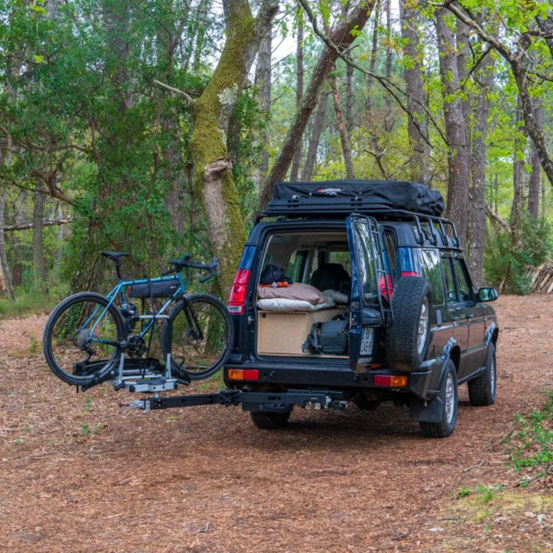 Un véhicule tout-terrain garé dans une forêt, avec un vélo sur un porte-vélos attaché à l'arrière, l'arrière du véhicule ouvert montrant des sacs et des équipements de camping.