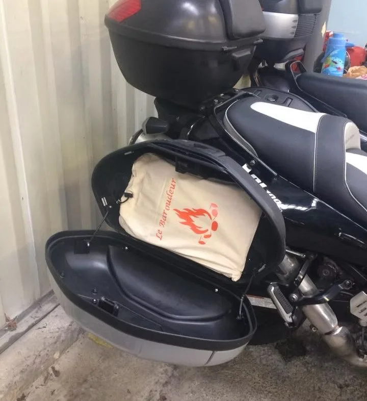 Une moto équipée d'une malle latérale ouverte contenant un sac beige avec un logo rouge d'une flamme et deux ronds, dans un garage, à l'intérieur, un barbecue "Le Baroudeur".