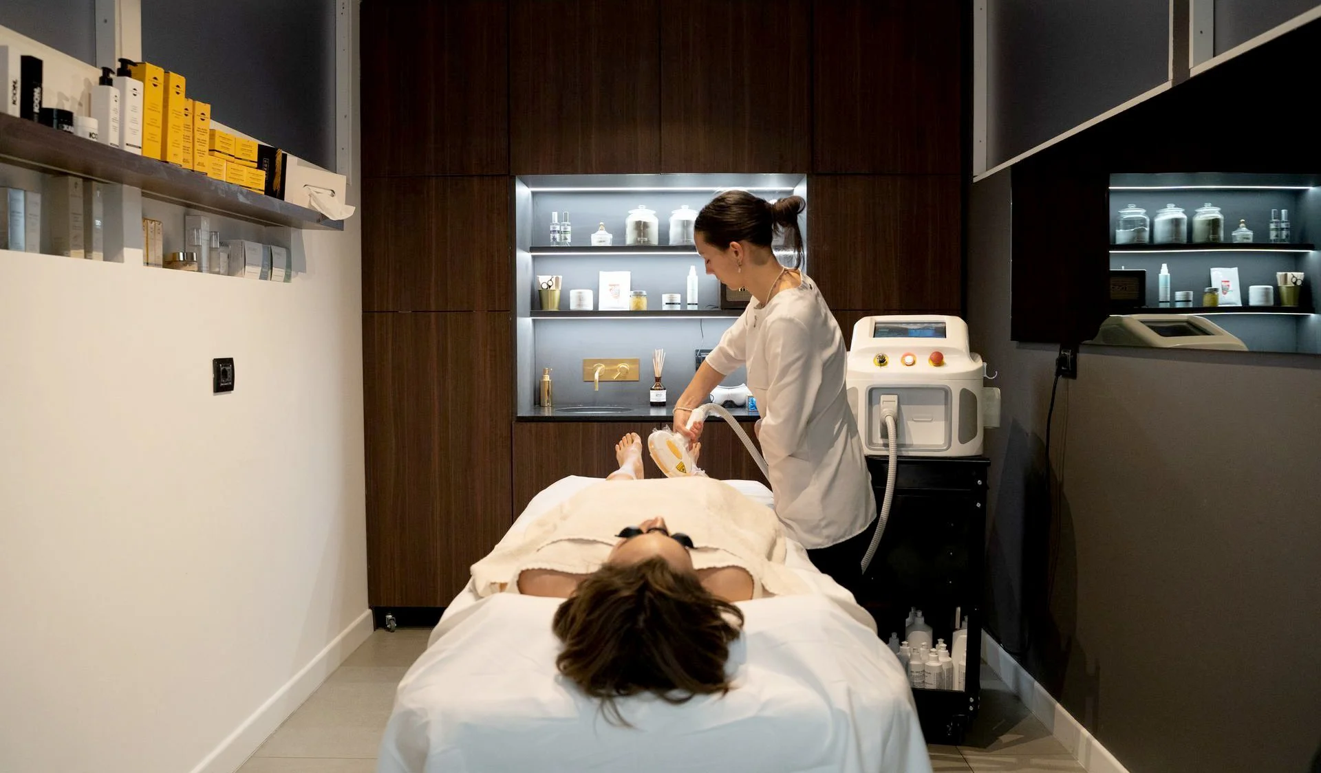 Una donna che riceve un trattamento estetico o dermatologico in un centro estetico, con una professionista che utilizza uno strumento su una cliente sdraiata su un lettino.
