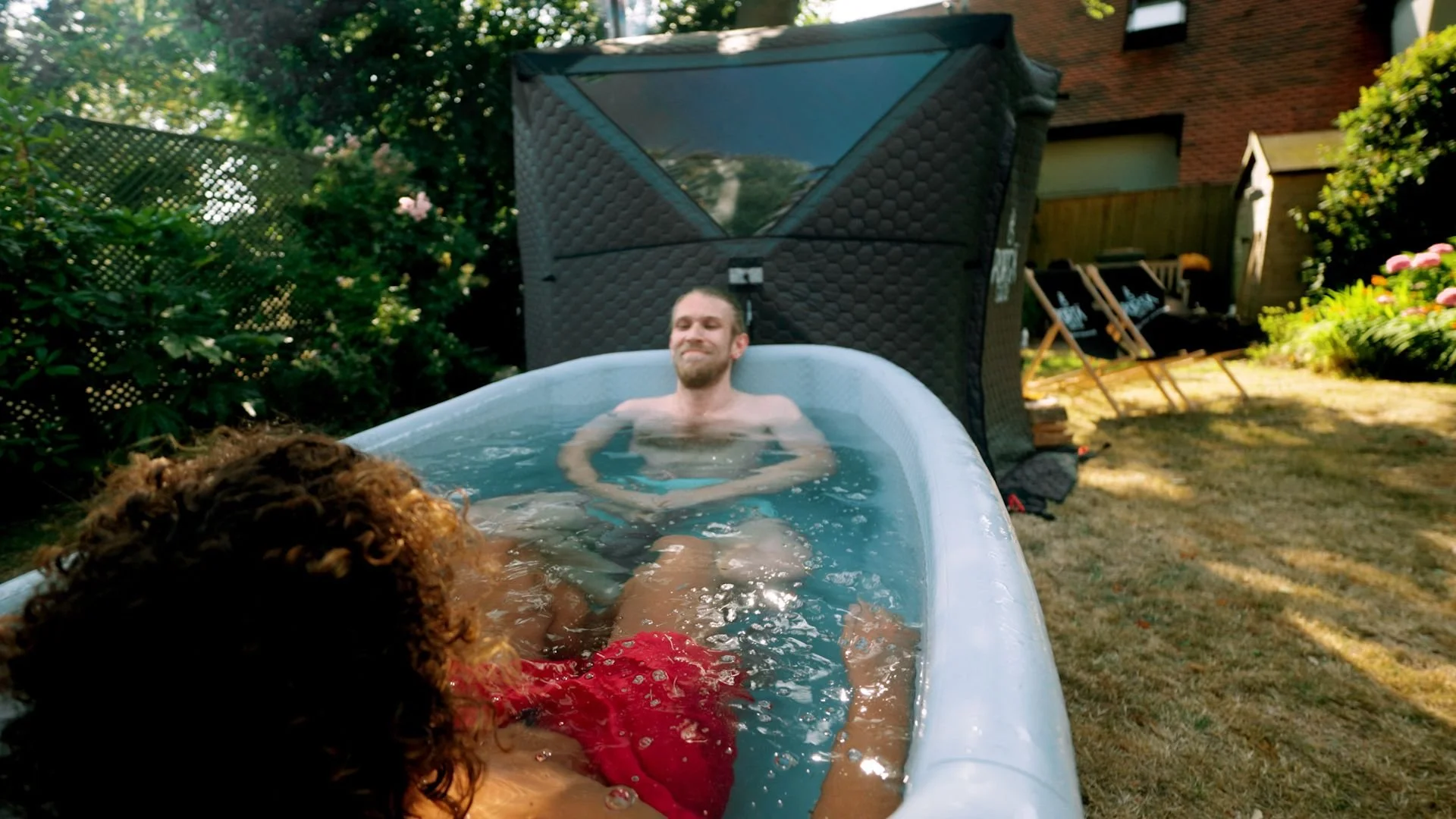 Two people relaxing in a cold plunge outdoors, with one man smiling and the other person’s head partially visible, surrounded by greenery and a backyard setting.