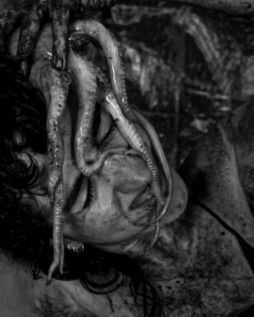 A black and white photo of a human face lying on the ground, with long hair and open eyes, partially covered octopus tentacles.