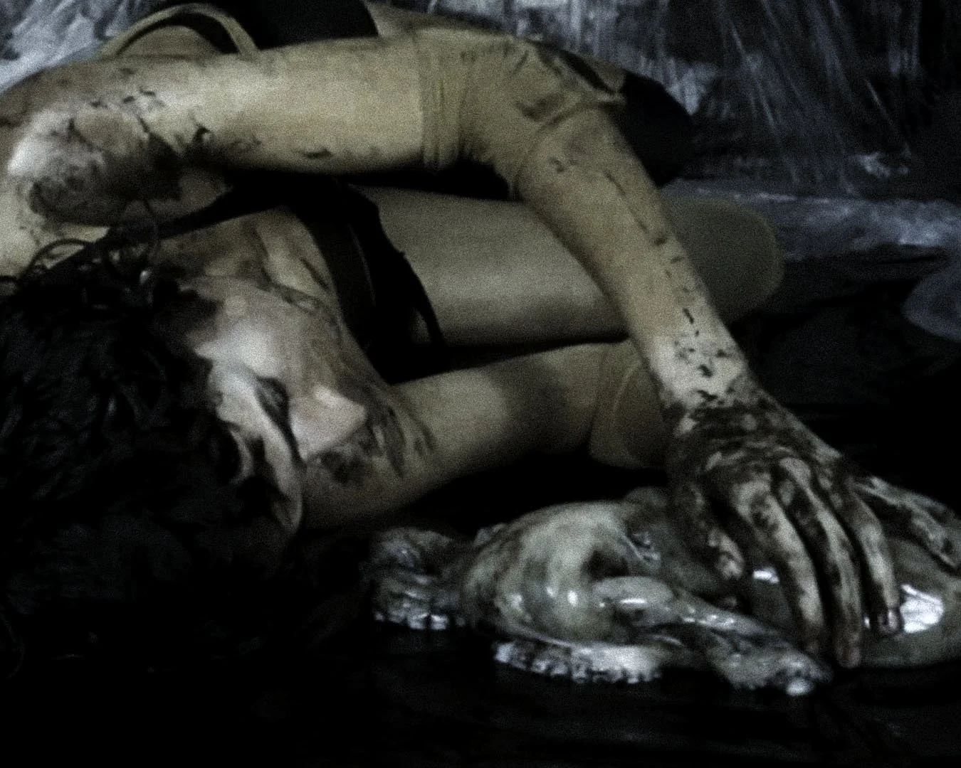 A person with long, dark, curly hair lying on the ground, holding a fish. The person is surrounded by dark, plastic-like material.