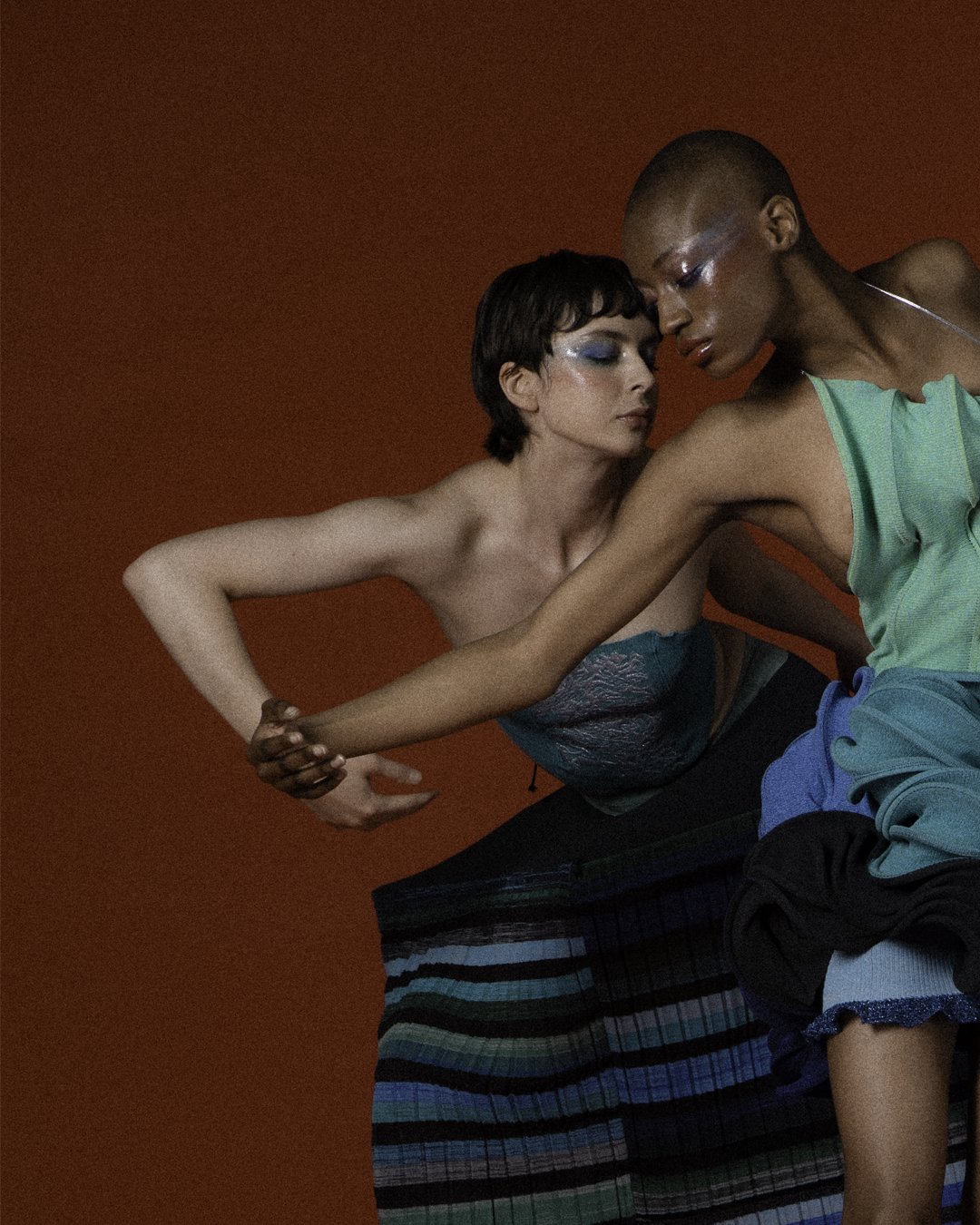 Two models with contrasting skin tones leaning into each other against a red background, with their eyes closed, touching hands, and dressed in colorful, fashionable clothing.