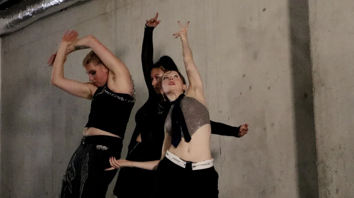 Three people dancing against a concrete wall, wearing fashionable black and metallic tops with expressive poses.