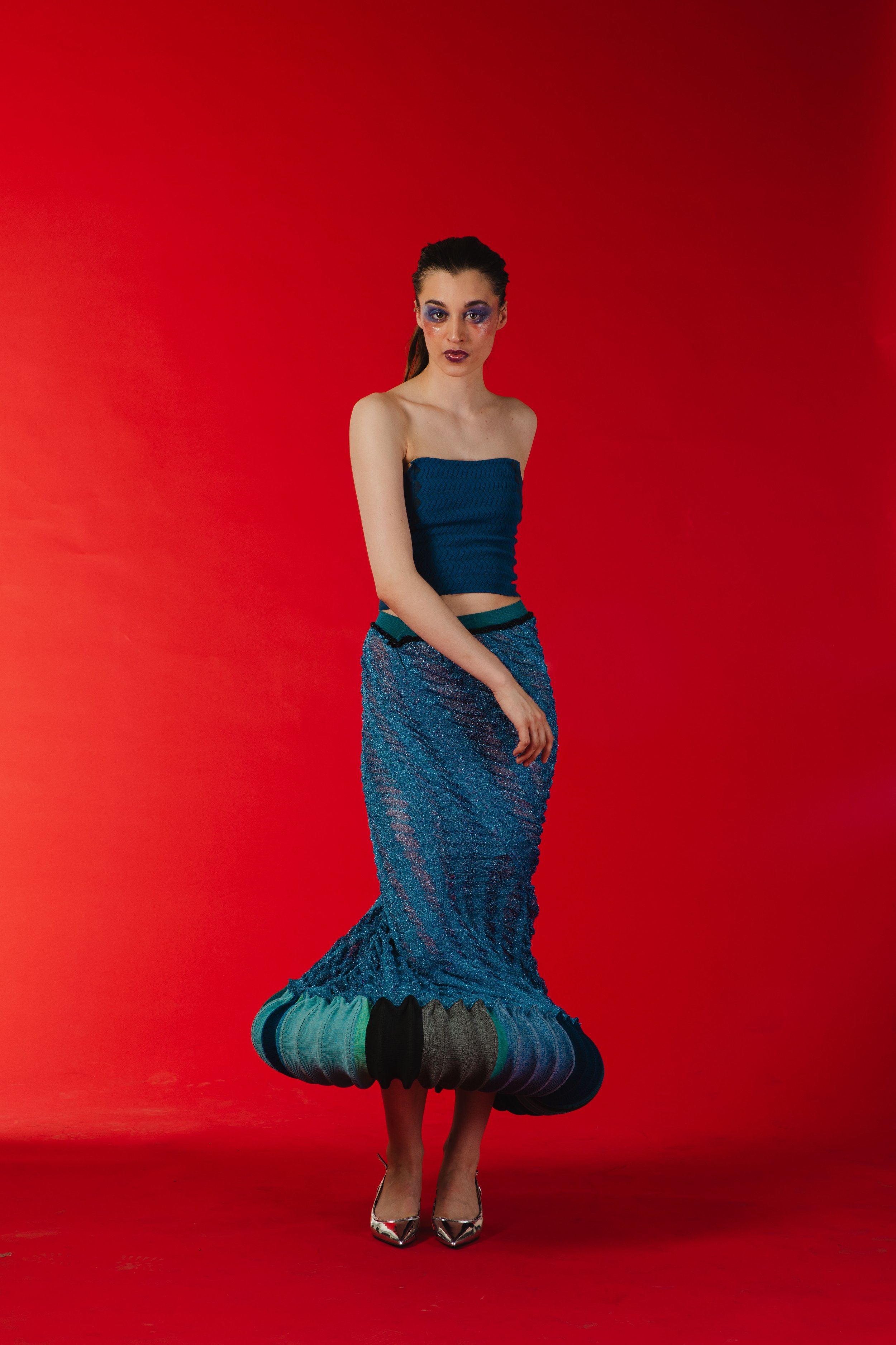 A woman wearing a strapless blue top and a long, textured, blue skirt with black and green accents, standing against a red background, with metallic high heels and dark makeup.