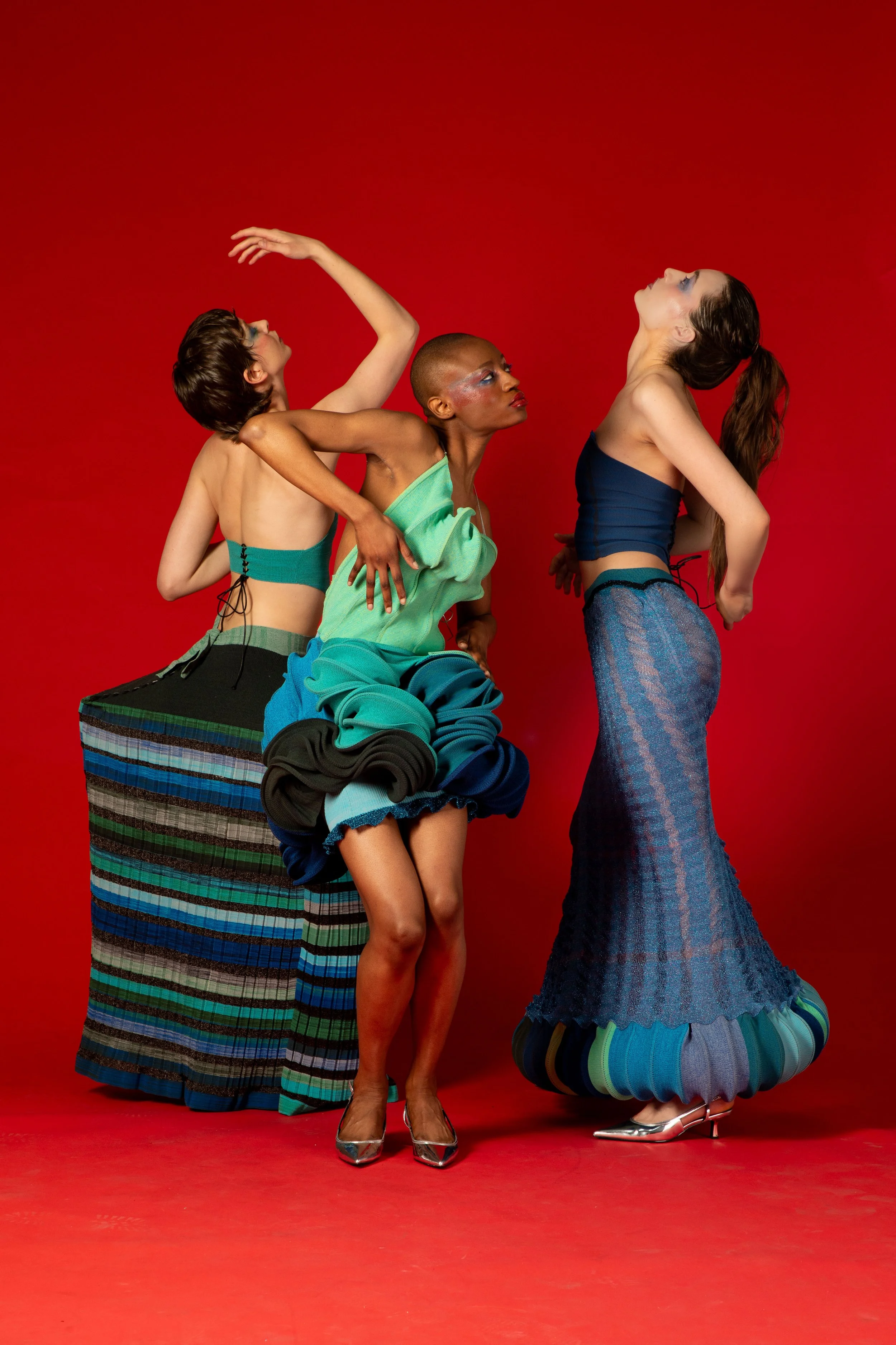 A diverse group of three women posing against a red background, dressed in colorful and expressive fashion with unique hairstyles and makeup.