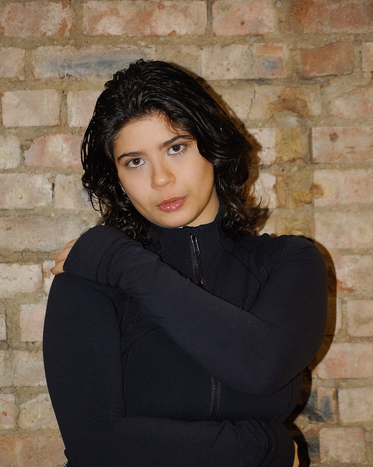 A woman with dark, shoulder-length curly hair, wearing a black zip-up jacket, posing against a brick wall background.