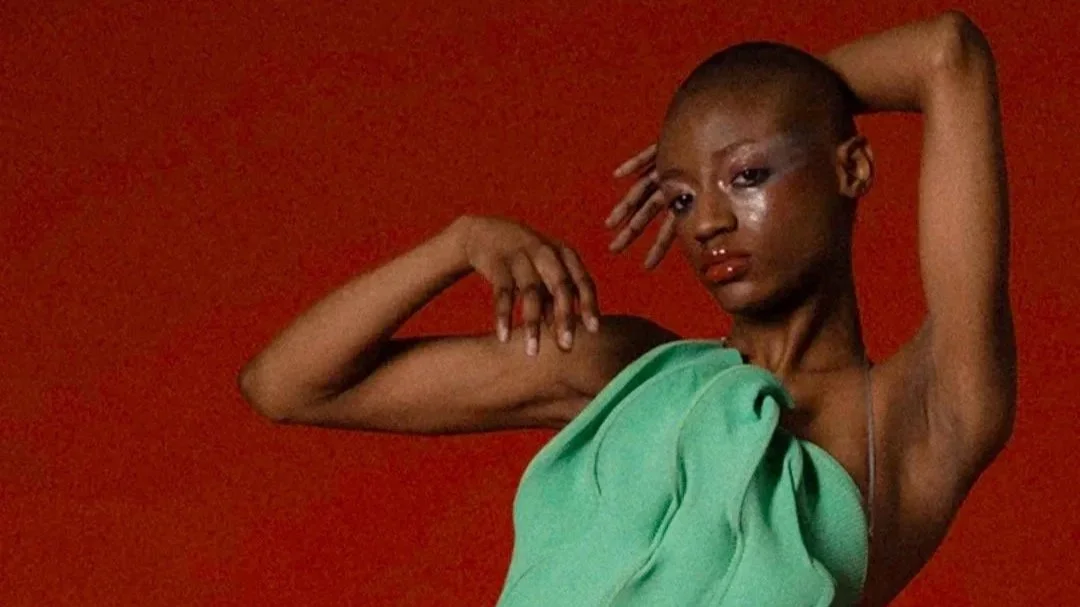 A person with buzzed hair posing with one arm flexed and the other behind their head, wearing a green garment against a red background.