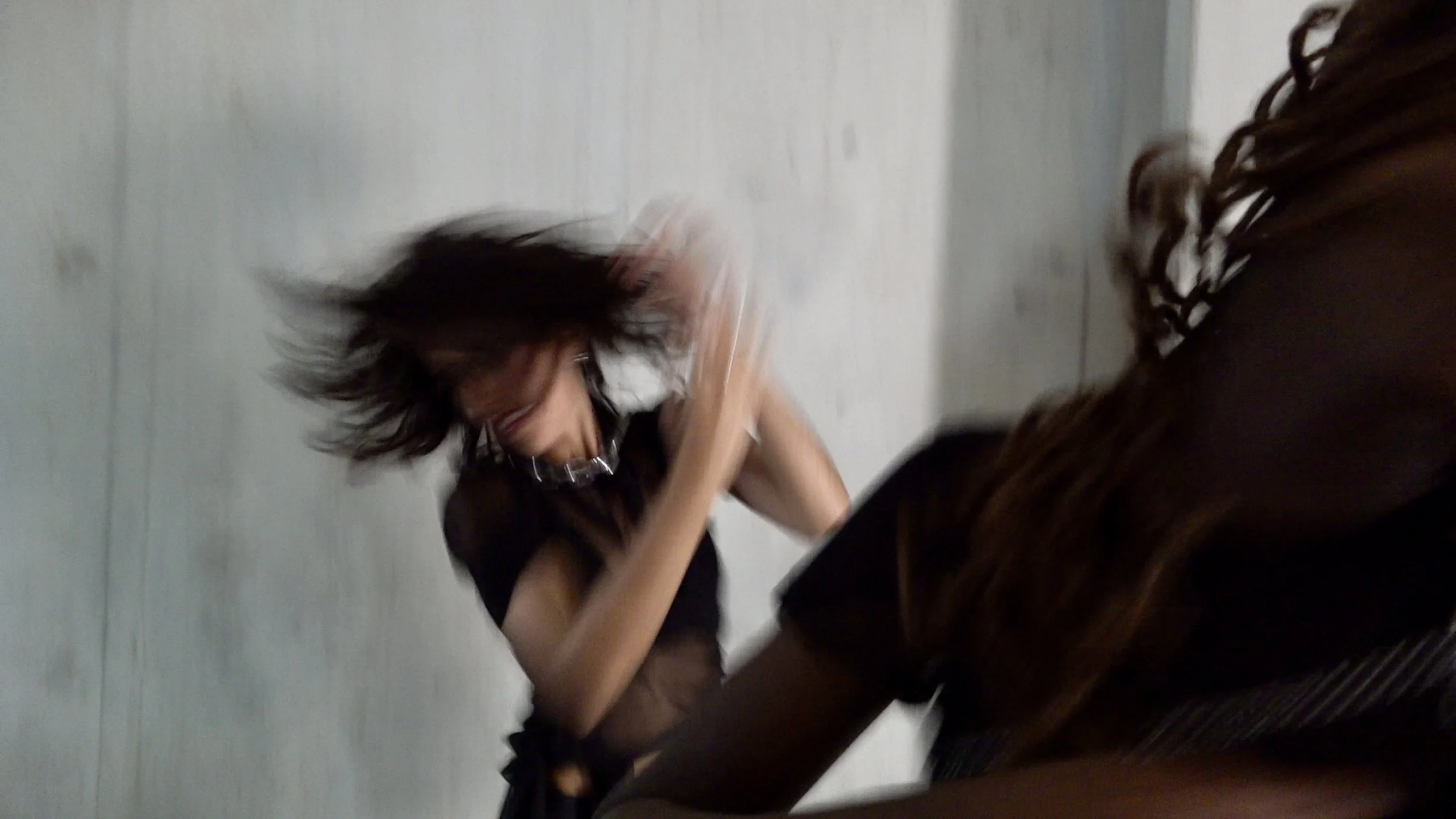 A woman with messy dark hair and chains appears to be in motion,  dancing against a plain gray wall.