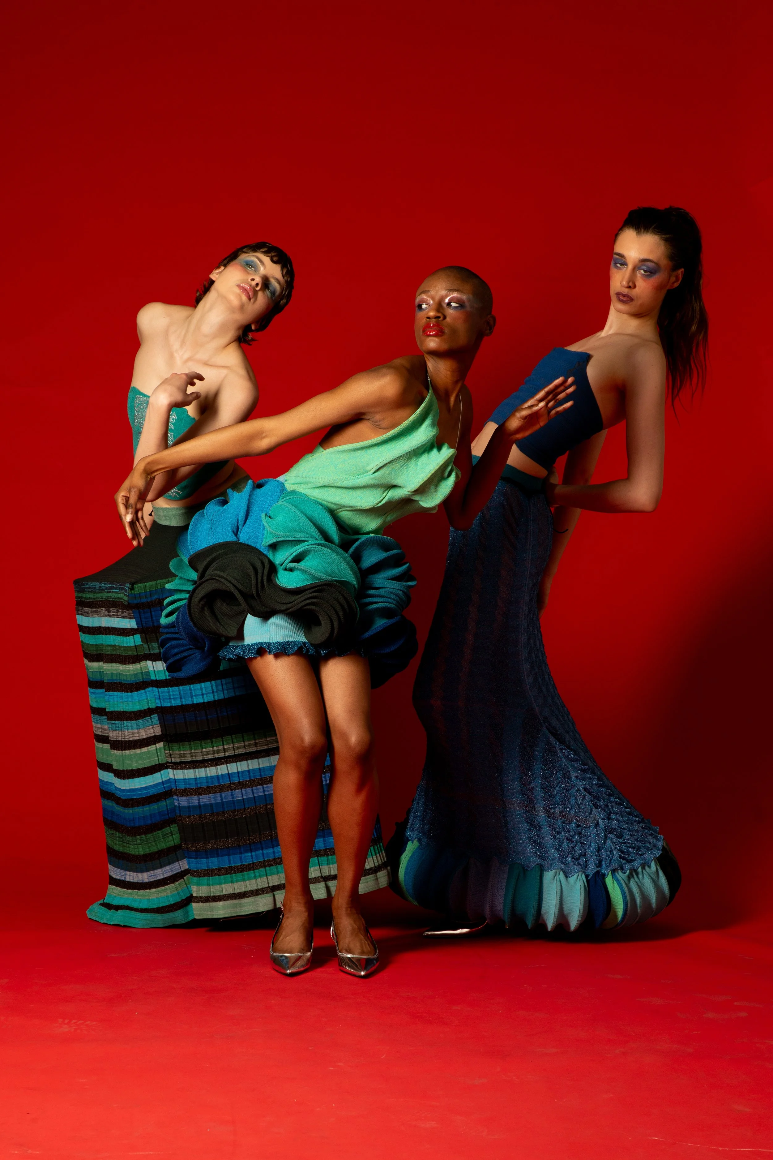 Three models with bold makeup and expressive poses against a red background, wearing eclectic and colorful fashion with a mix of textures and patterns.