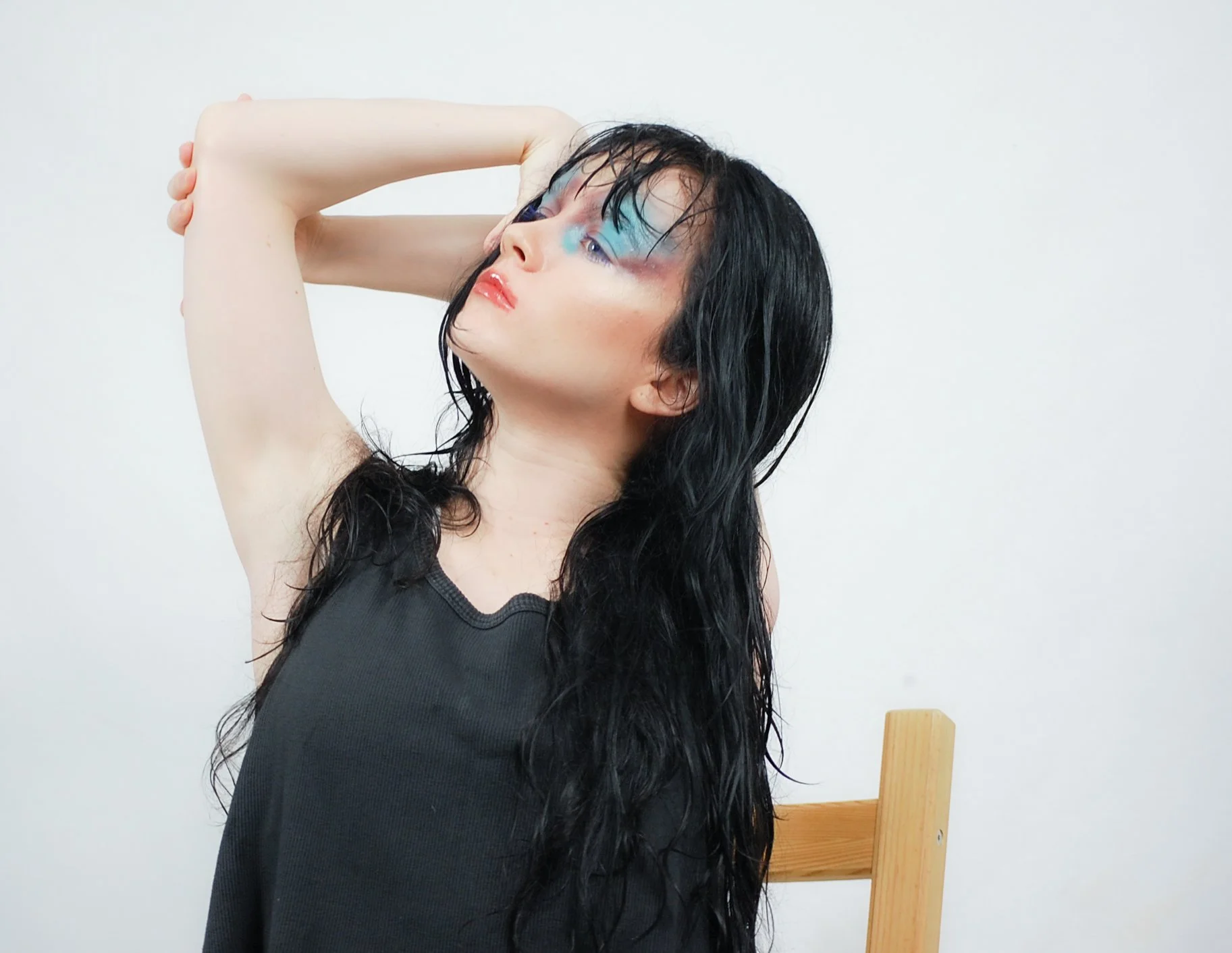 A woman with long black hair and colorful makeup, wearing a sleeveless black top, posing with one arm raised and hand on her head, standing next to a wooden chair against a plain white wall.