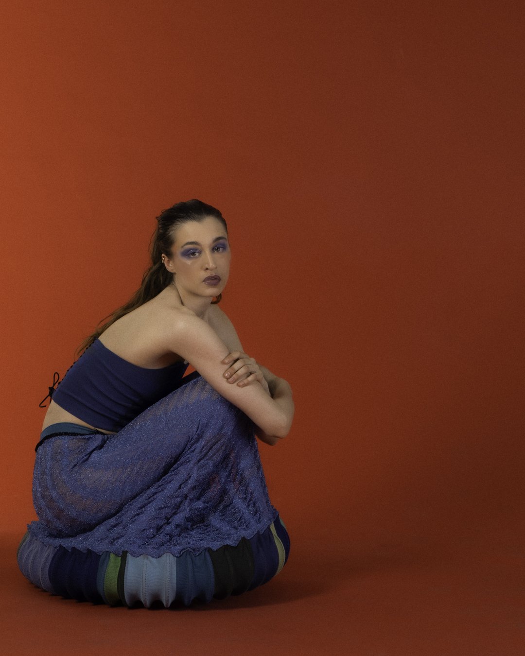 A young woman with long brown hair kneeling against a red background. She is wearing a blue tank top and a blue skirt, with her arms crossed over her knees and looking at the camera.