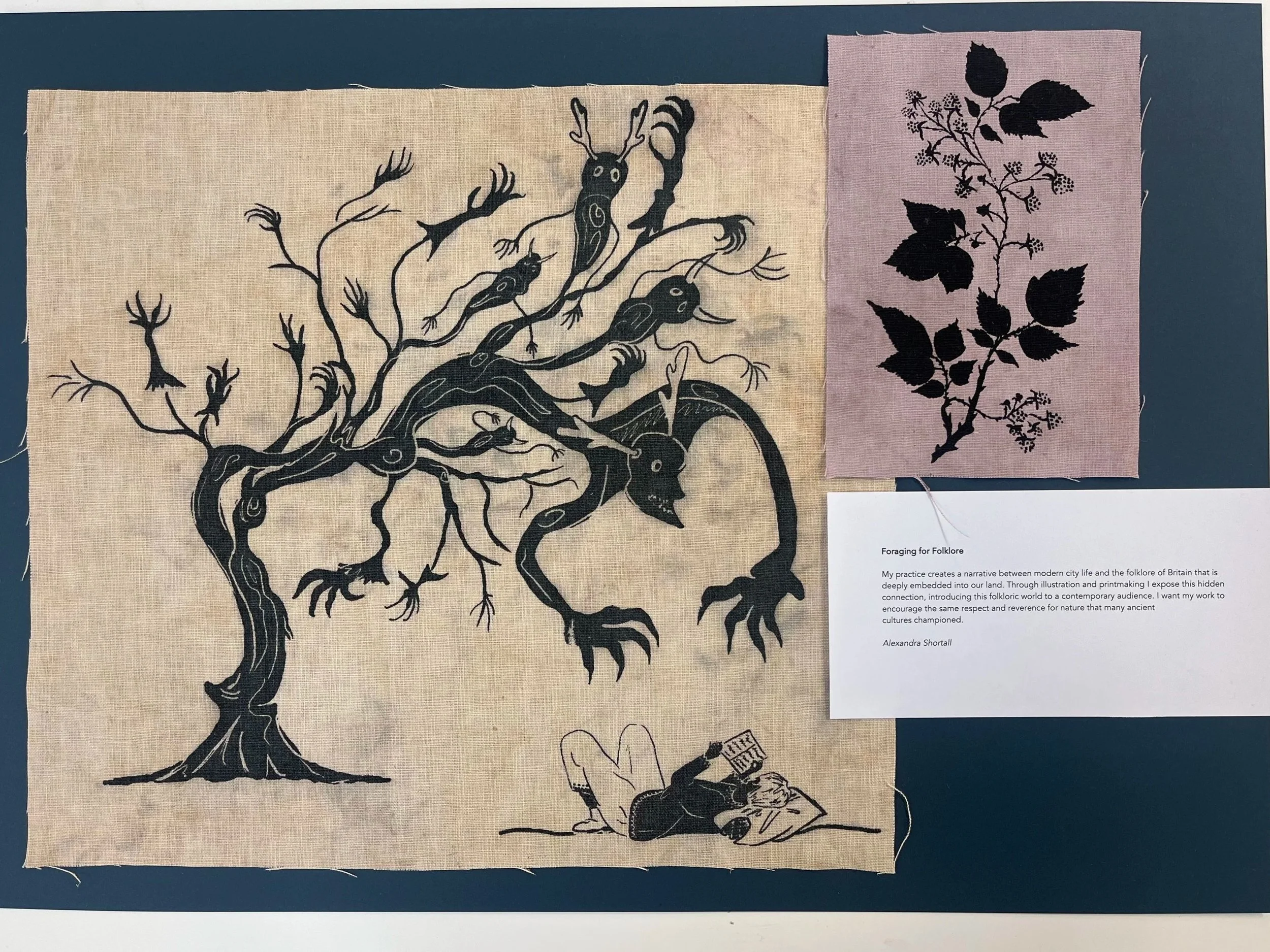 A black silhouette of a large, leafless tree with twisting branches on a beige fabric background, with several bird and animal figures integrated into the tree's design. To the right is a smaller pink fabric piece with a black floral pattern of leaves and branches. Below is a printed description discussing folklore, with mention of Alexandra Shortall.