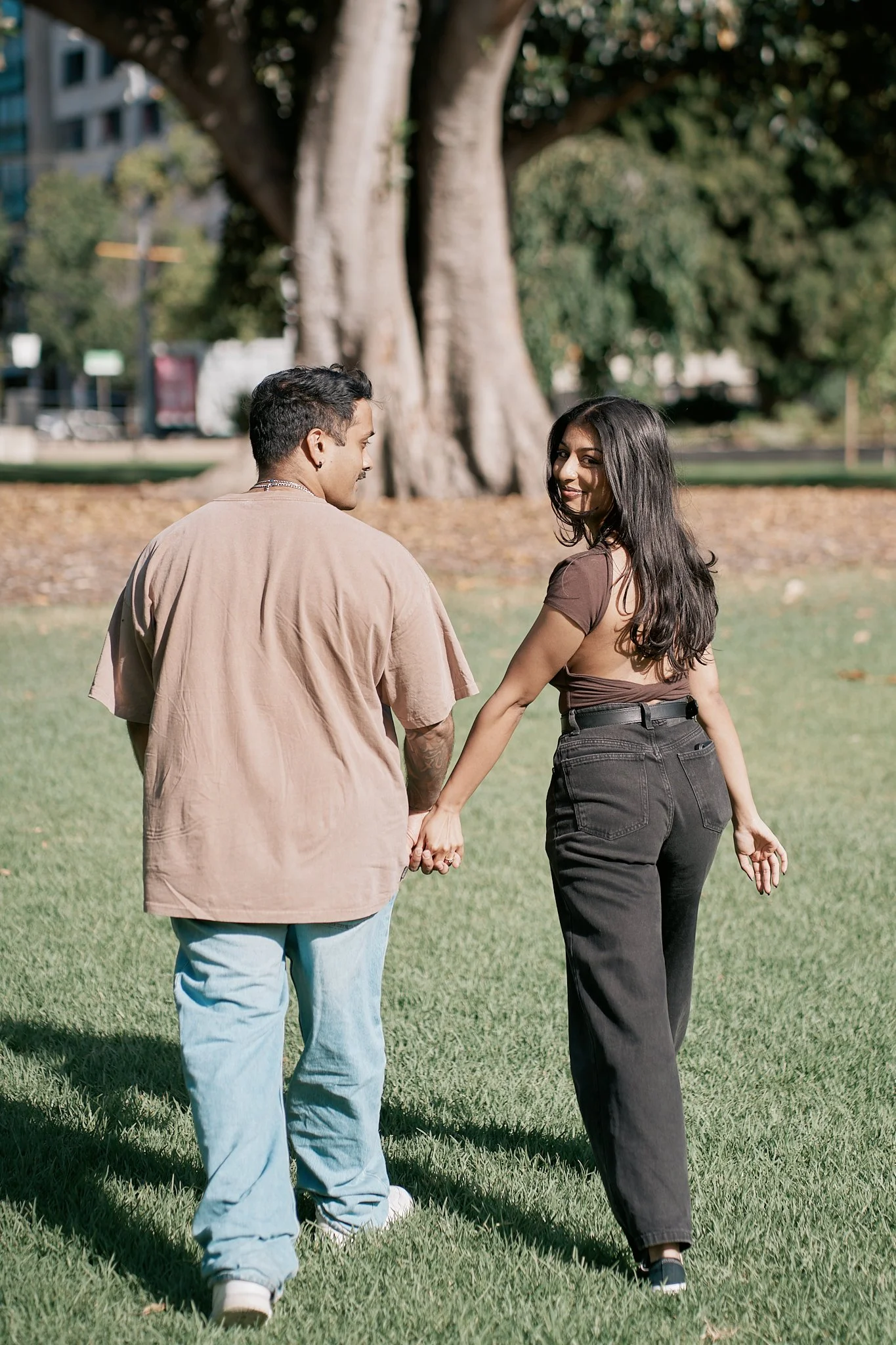 A couple holding hands walks through a park with green grass and large trees, smiling at each other.