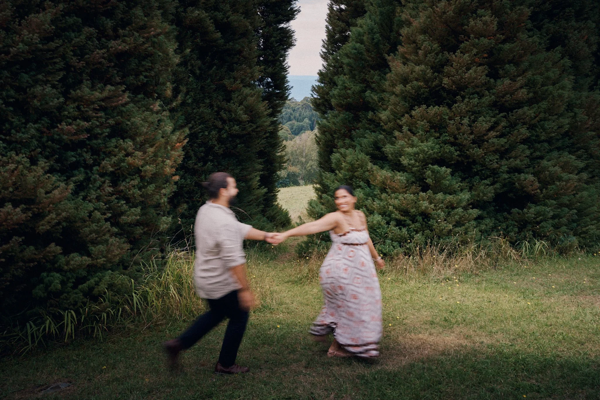 Two women holding hands and dancing outdoors on a grassy clearing surrounded by tall pine trees, with a partly cloudy sky and distant horizon in the background.