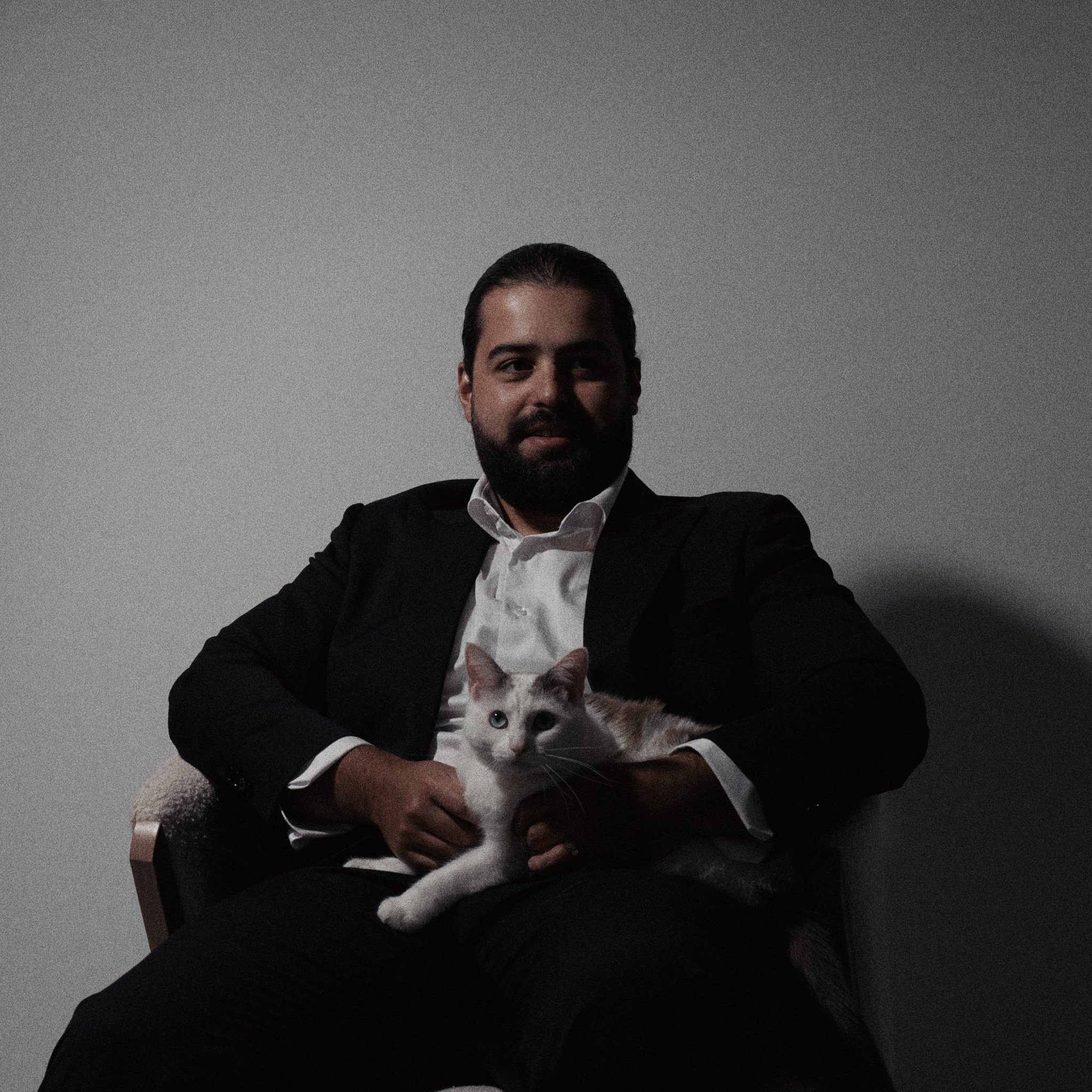 A man in a black suit with a white shirt sitting on a chair, holding a white cat with orange and black markings, in a dimly lit room with a plain gray wall background.