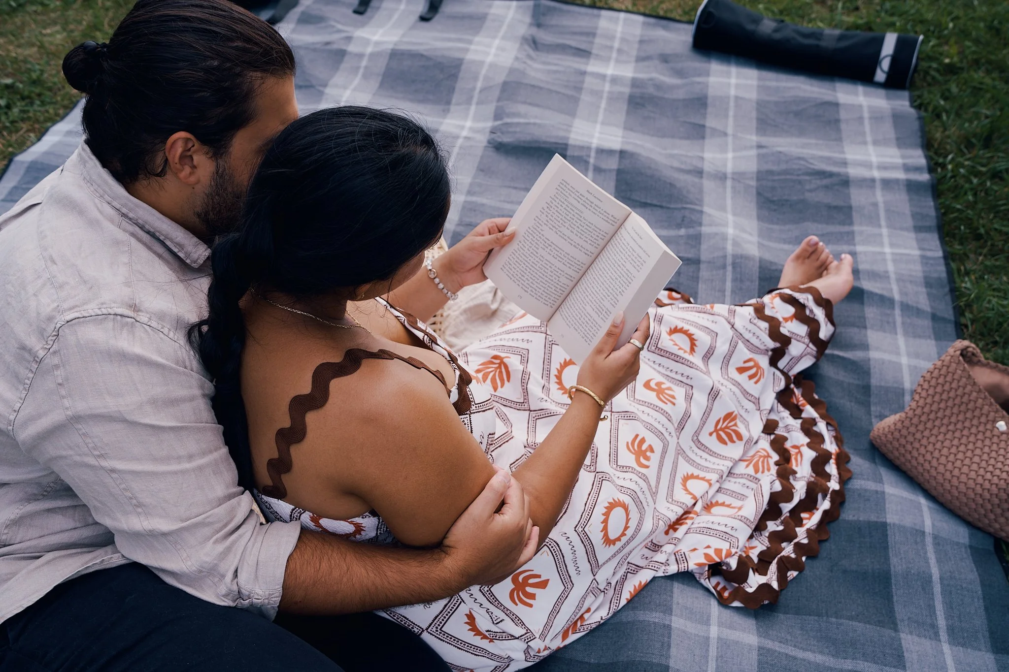 A couple sitting on a plaid blanket outdoors, with the woman reading a book. The woman wears a patterned dress with brown accents, and the man lounges behind her with his arms around her.