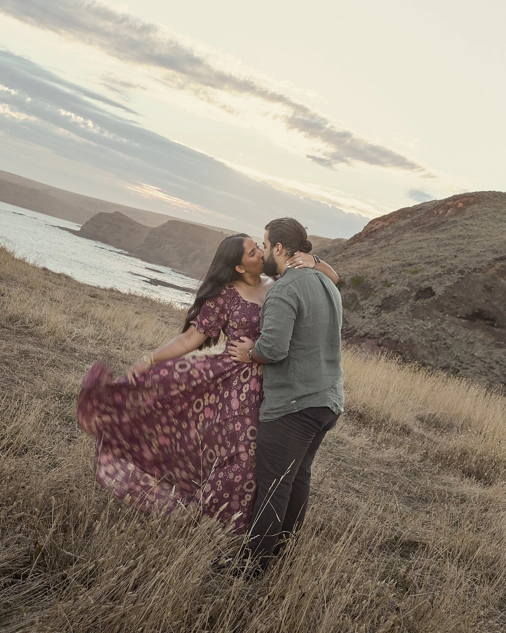 A couple sharing a kiss in a grassy field near the coast during sunset, with cliffs in the background.
