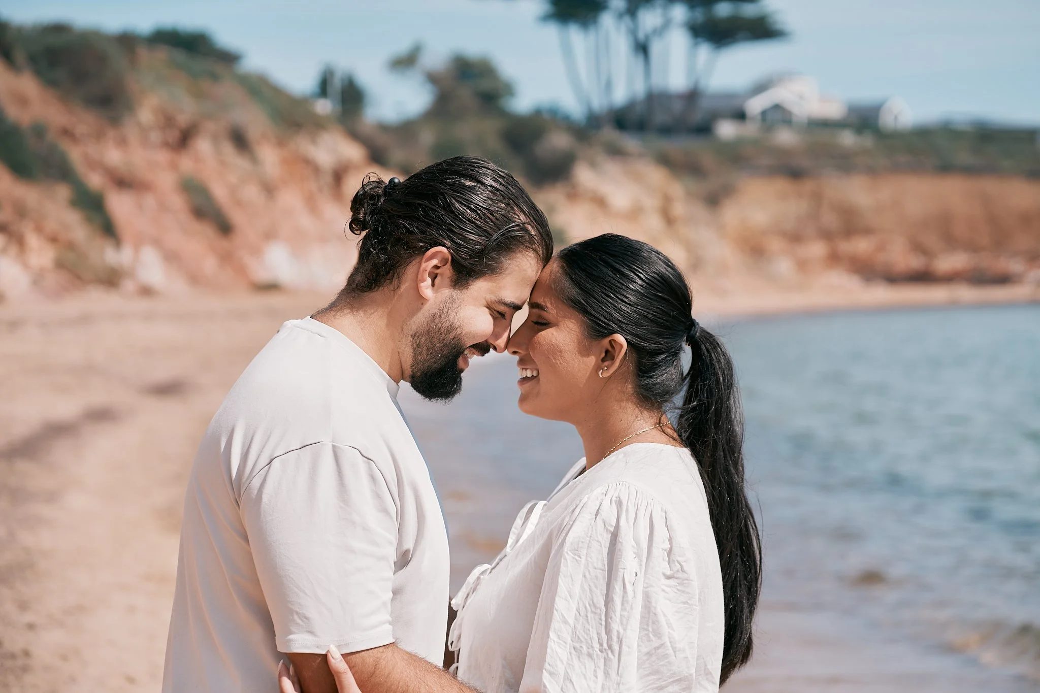 A couple with their foreheads touching, smiling, standing on a beach with cliffs and trees in the background.