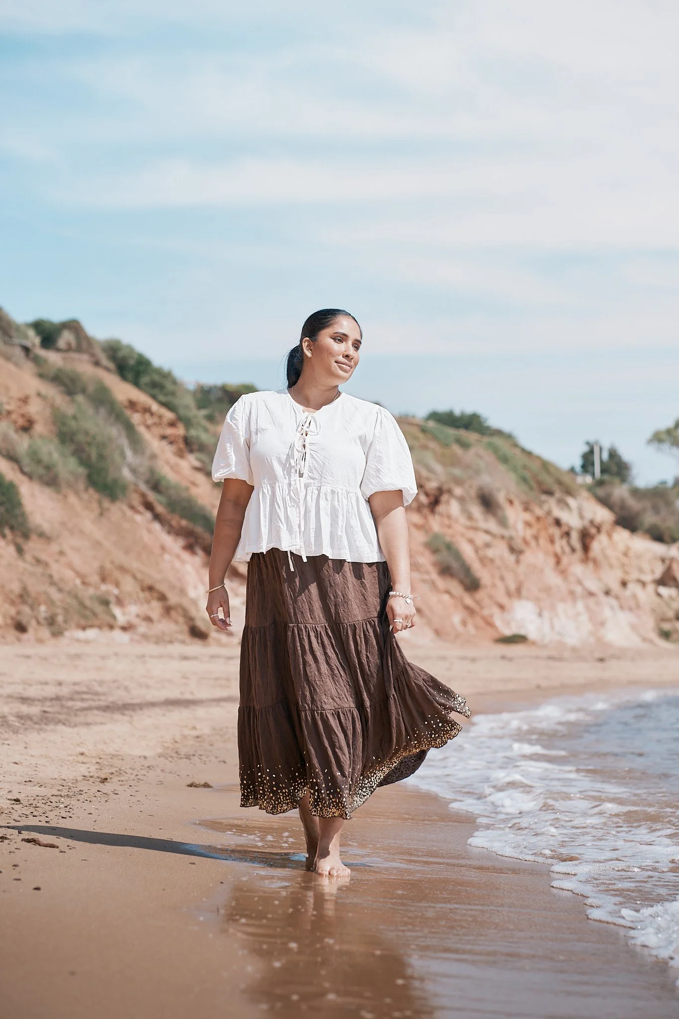 Woman walking barefoot on the beach with rocky cliffs in the background, wearing a white blouse and a long brown skirt.