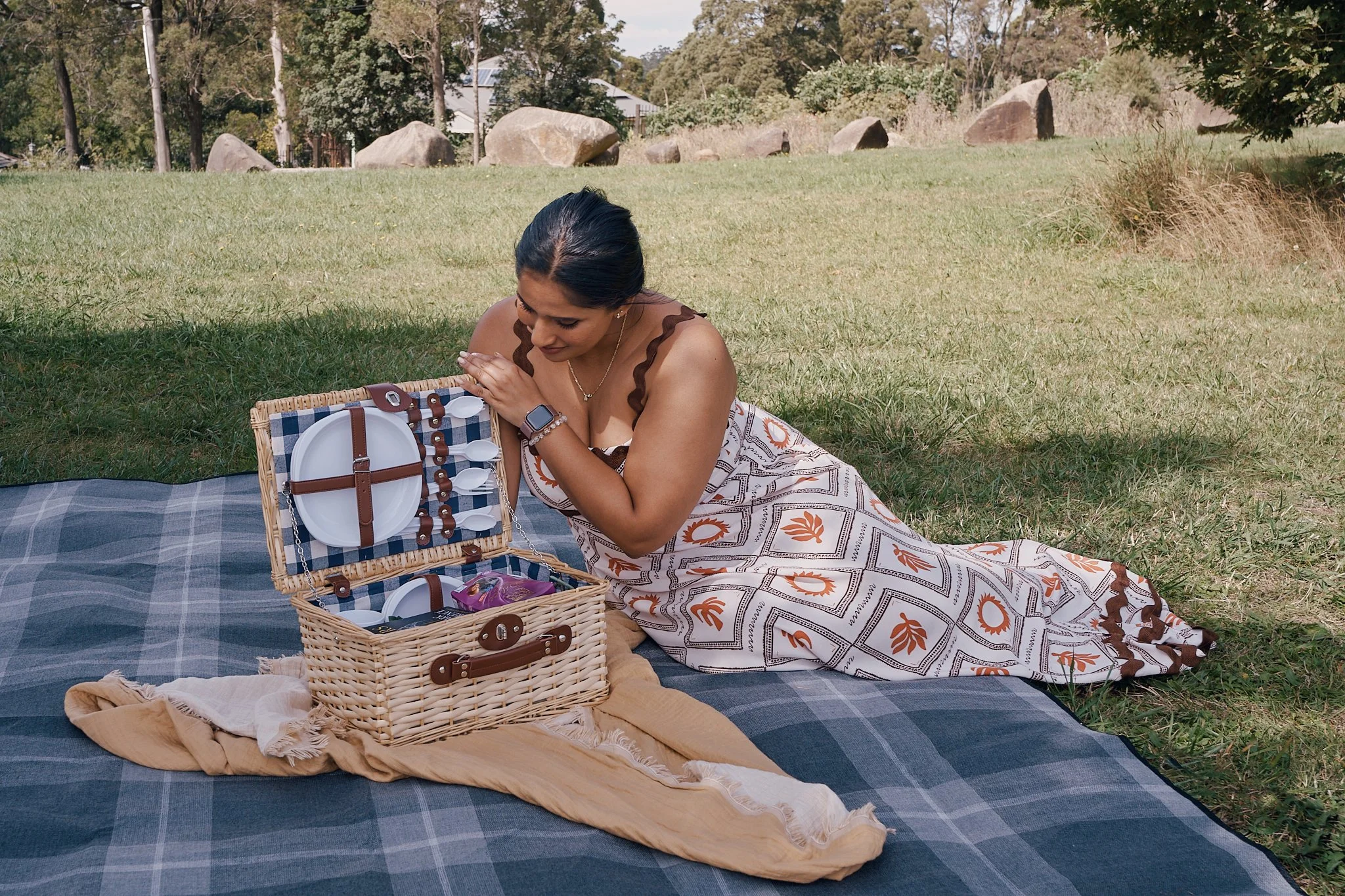 A woman in a white and orange patterned dress sitting on a blue and gray blanket in a park, looking into a picnic basket with picnic plates, utensils, and snacks inside.