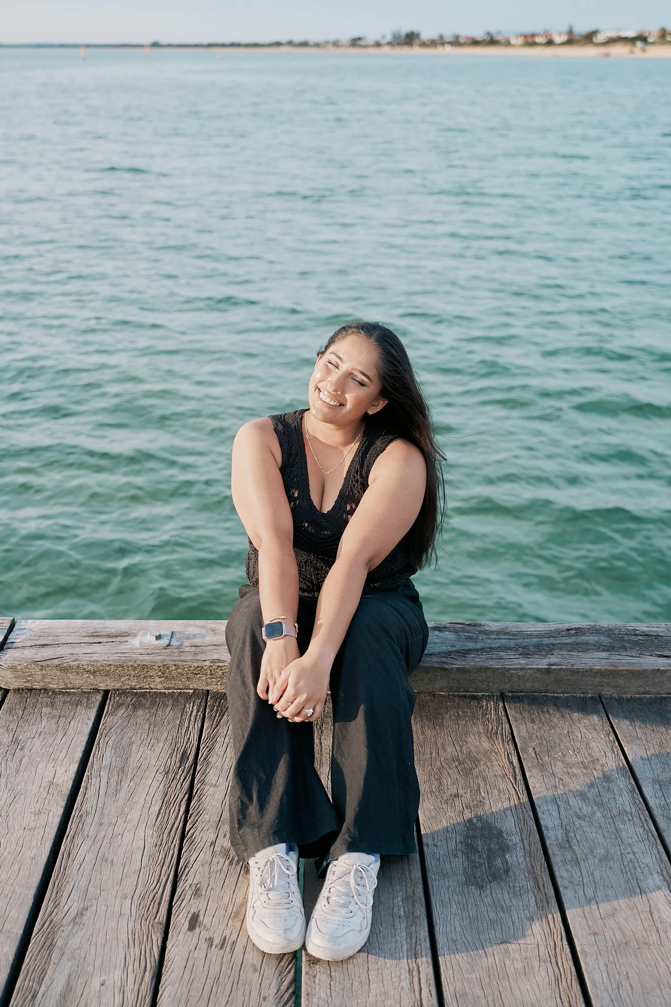 A woman sitting on a wooden dock by the water, smiling with her eyes closed.