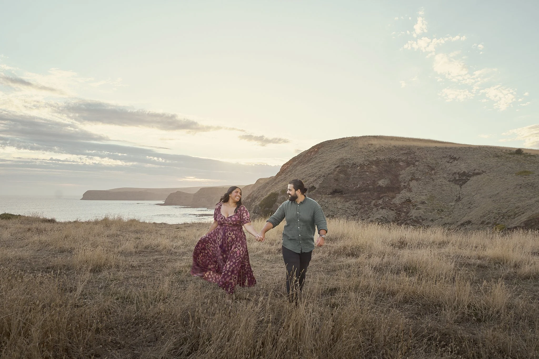 A couple walking hand in hand through a grassy field at sunset, with cliffs and ocean in the background.