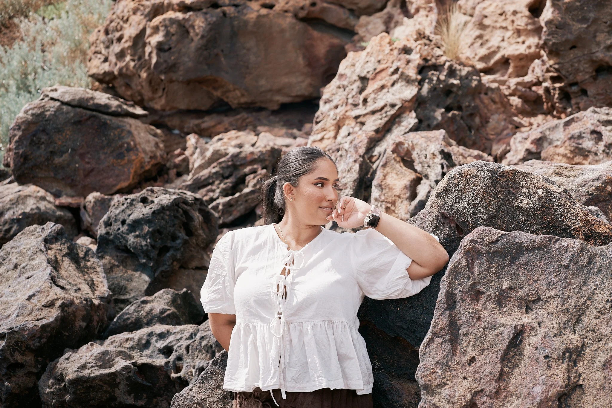 A woman with dark hair tied back, wearing a white blouse with a front tie and brown pants, leaning against large, dark-colored rocks outdoors during daytime.