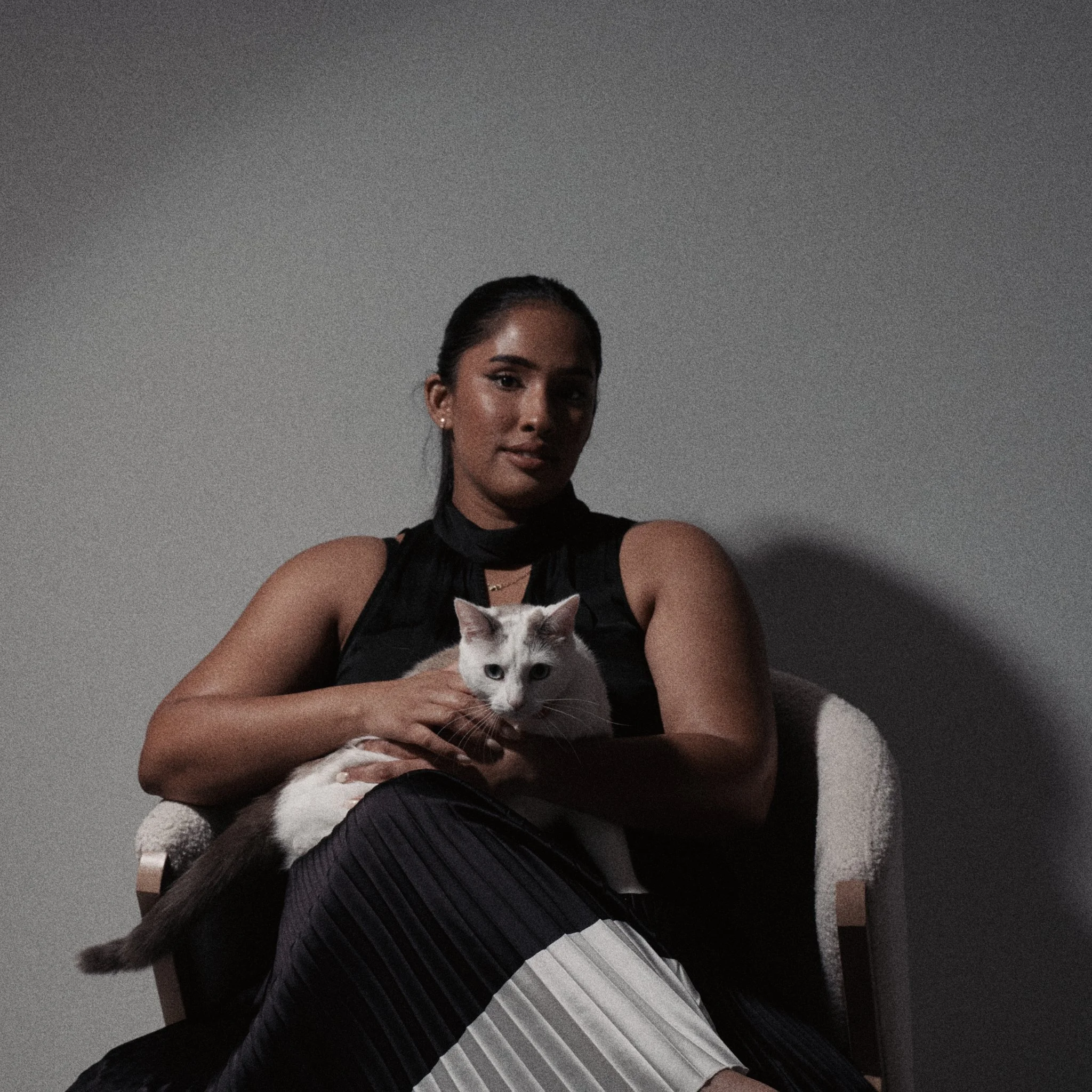 A woman sitting on a chair holding a white cat with blue eyes against a plain gray wall.