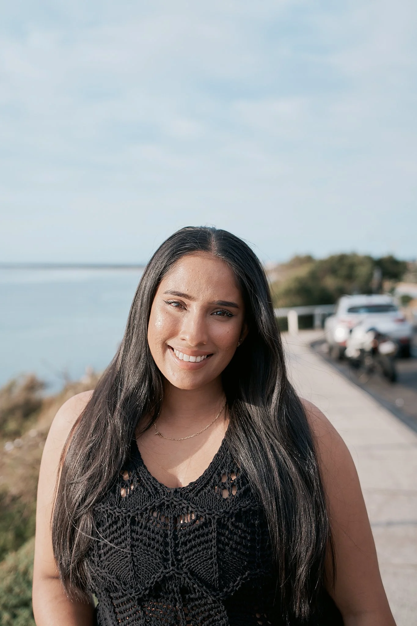 A young woman with long dark hair smiling outdoors near the water and a parking lot.
