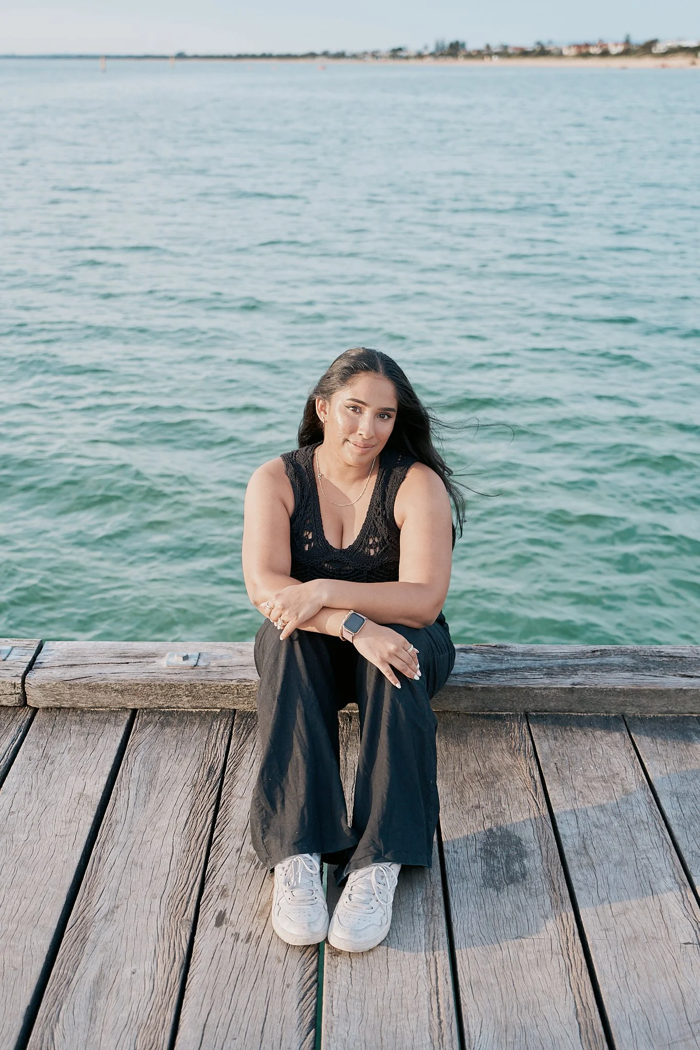 A woman sitting on a wooden dock by the water, smiling at the camera, with the ocean and distant shoreline in the background.