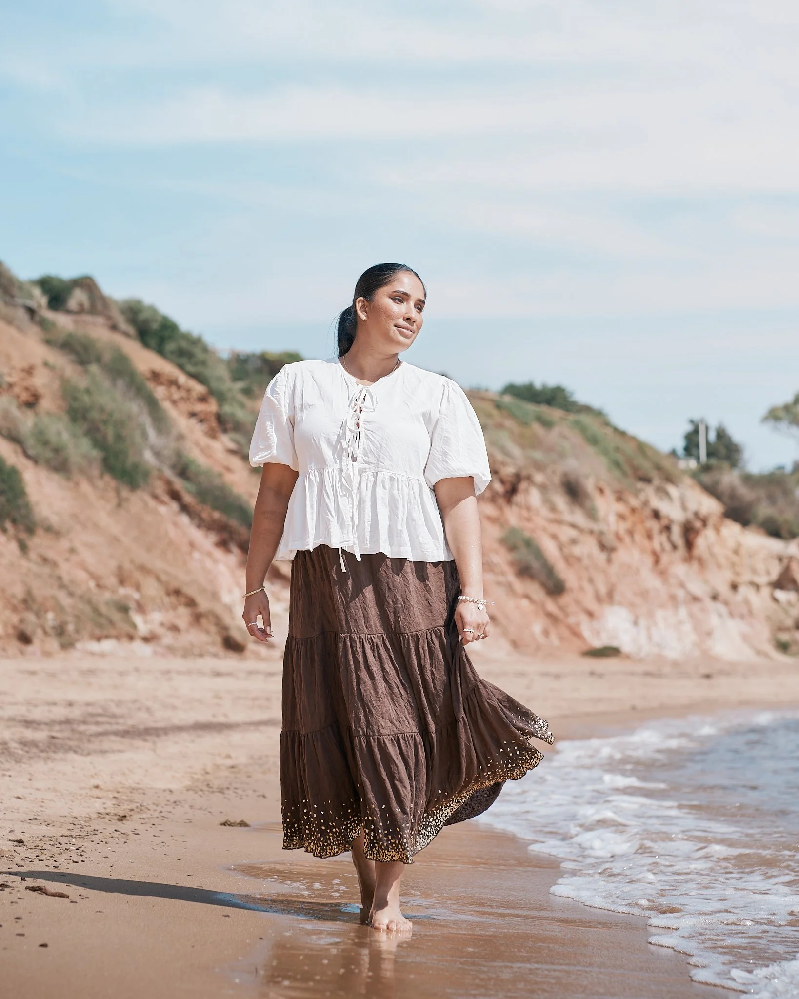 A woman walking barefoot on the beach, wearing a white blouse and a brown maxi skirt, with sandy cliffs and a partly cloudy sky in the background.