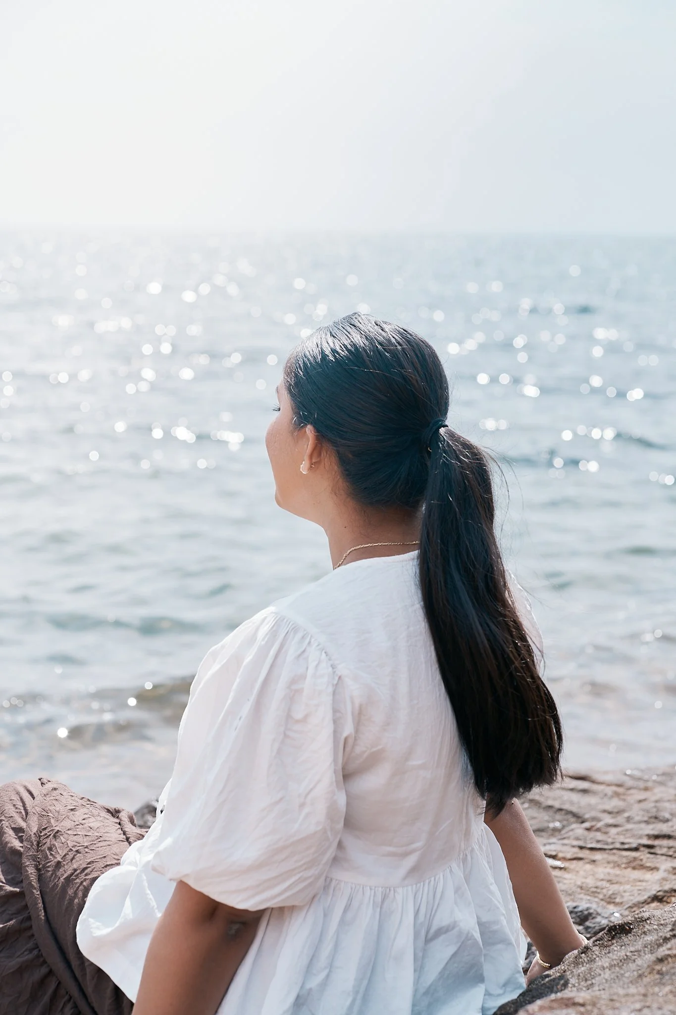 A woman with long dark hair in a ponytail, wearing a white blouse, sitting by the ocean shore, looking out at the water.