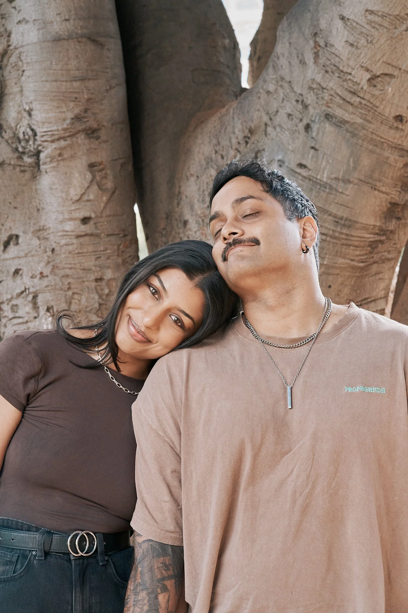 A man and woman sharing a moment with the woman resting her head on the man's shoulder in front of large tree trunks.