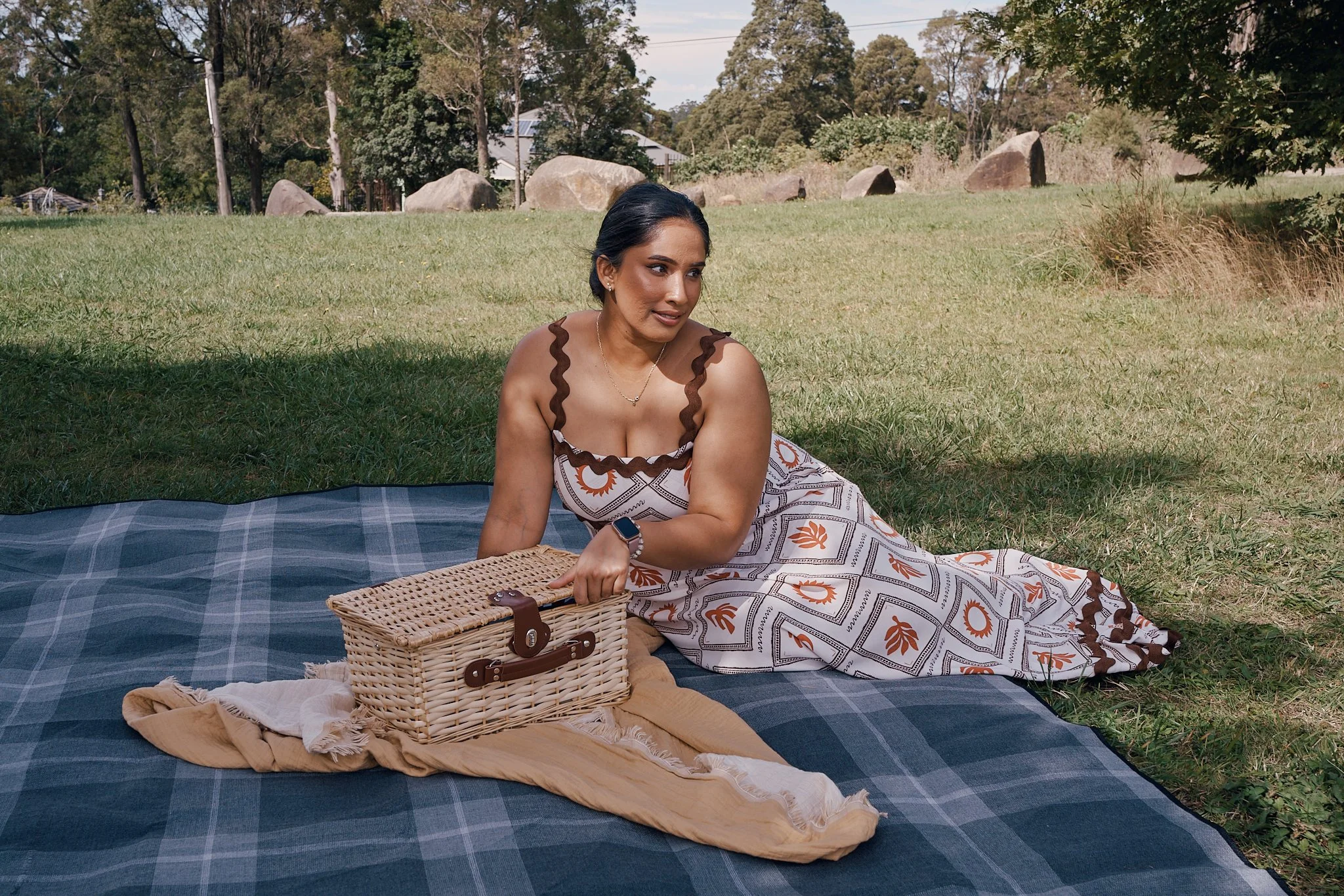 A woman in a patterned dress is sitting on a blue and black checkered blanket on the grass in a park, with a wicker picnic basket nearby.
