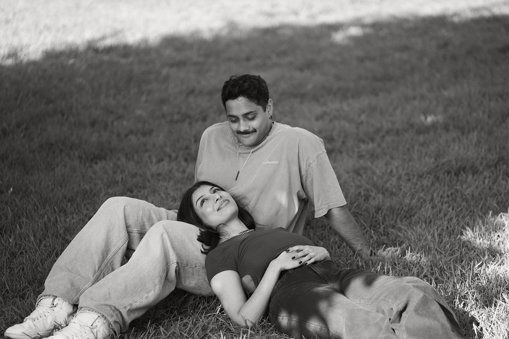 A young couple relaxes on the grass outdoors, with the woman lying on her back and the man sitting behind her, both smiling and enjoying each other's company.