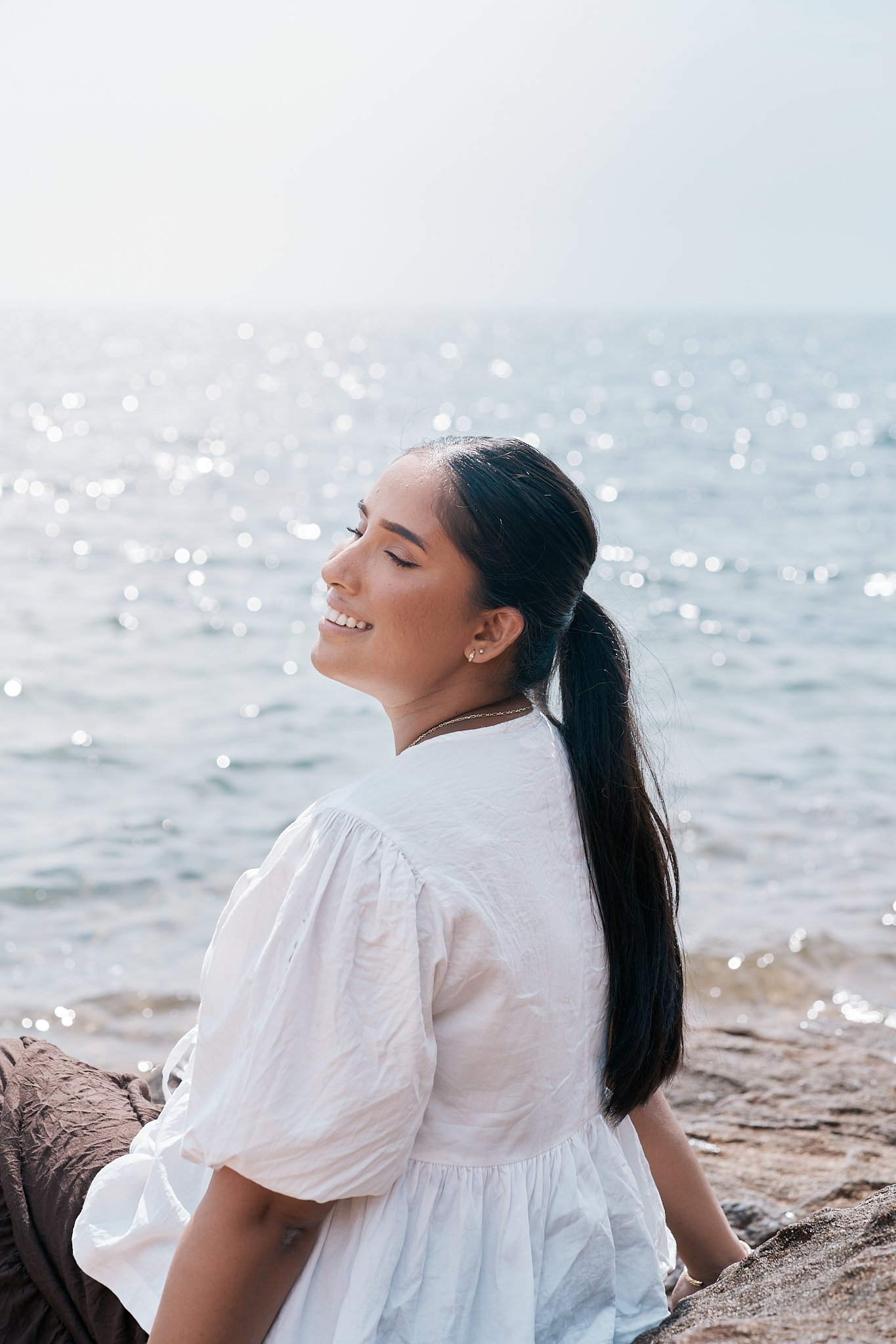 A woman with long black hair sitting on rocks by the water, smiling with her eyes closed, enjoying the sunny day at the beach.