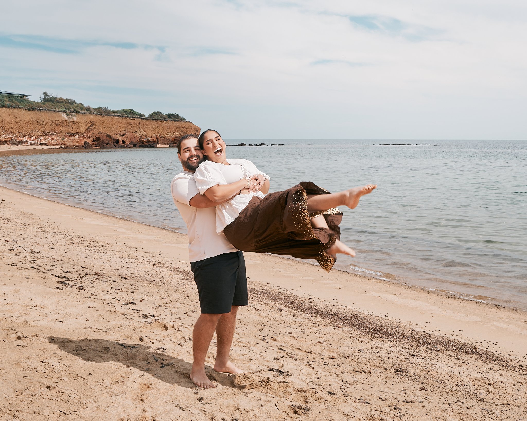 A man is holding a woman in his arms on a beach, both are smiling and laughing. The woman is laying back with her legs extended outward, wearing a white top and a brown skirt, while the man is wearing a white T-shirt and black shorts. The beach has sand, and the ocean and partly cloudy sky are in the background.
