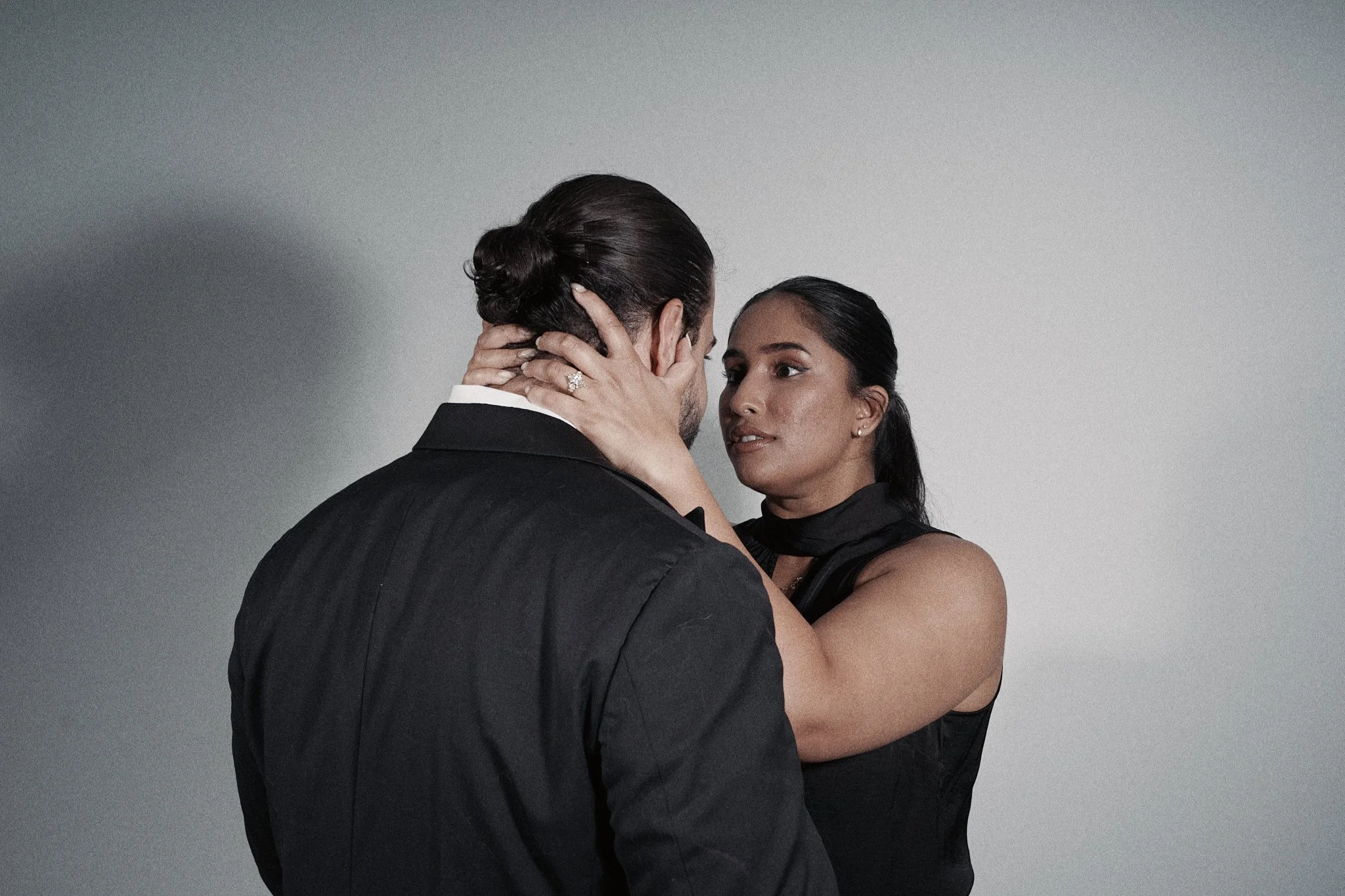 A woman and a man in formal attire face each other closely, with the woman holding the man's face gently and looking into his eyes against a plain background.