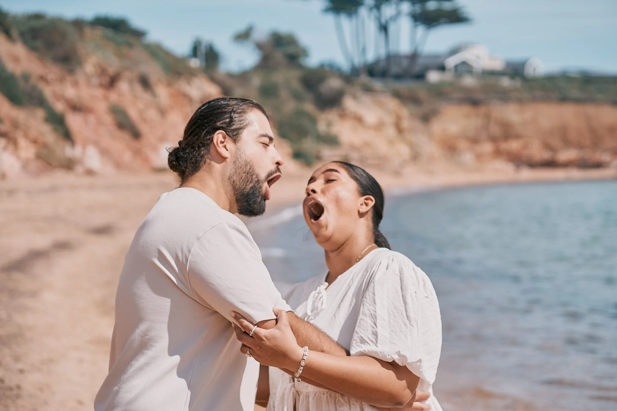 A man and woman on a beach yawn or pretend to yawn, with ocean and cliffs in the background.