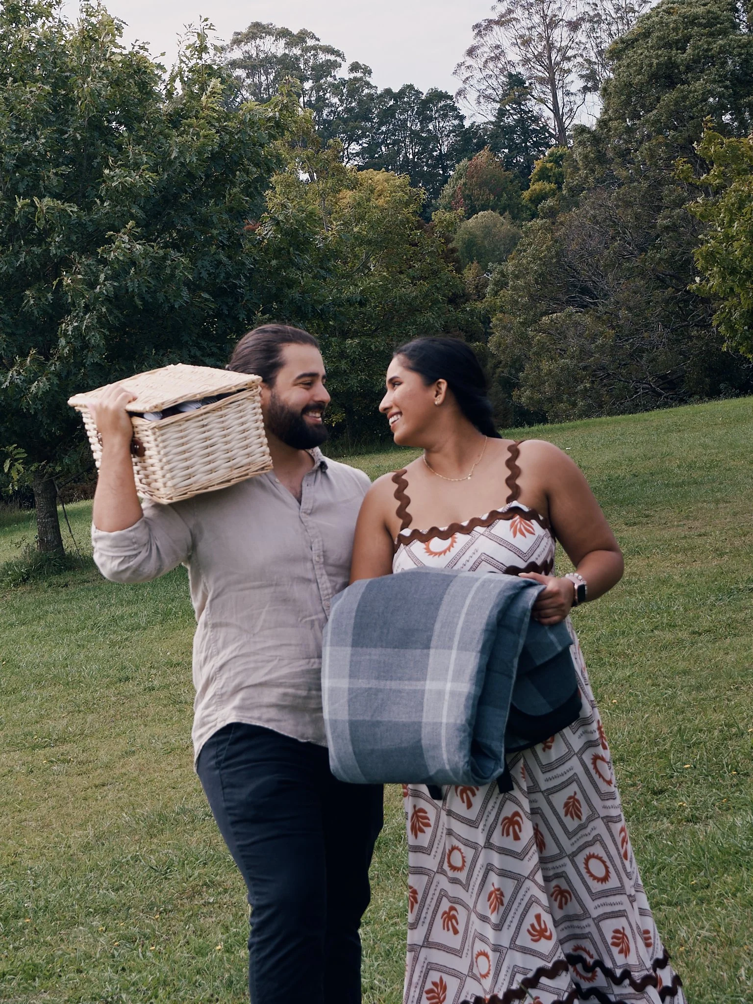 A man and woman smiling at each other outdoors during a picnic, with the man carrying a picnic basket and the woman holding a blanket, standing on a grassy area with trees in the background.
