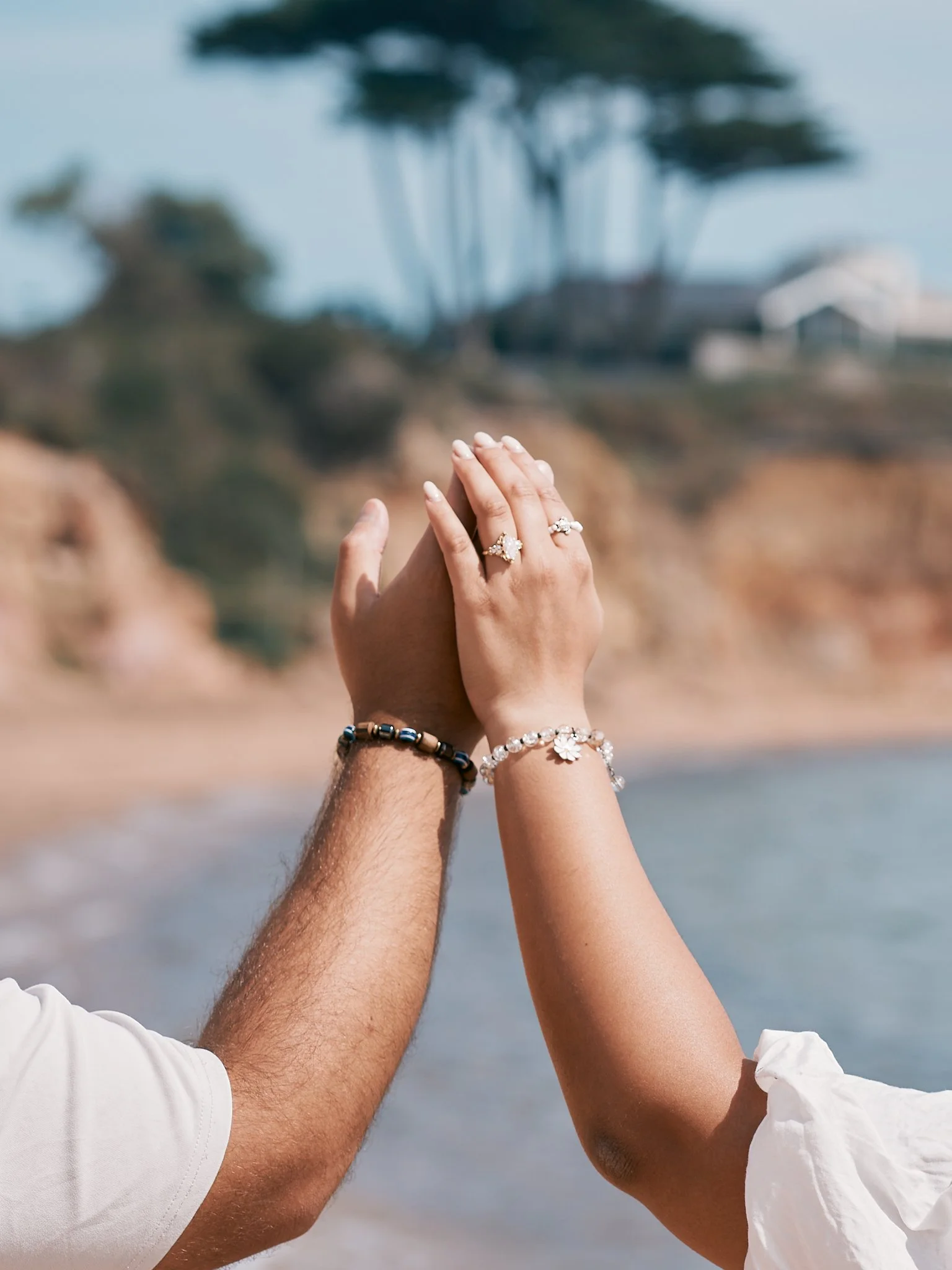 Close-up of a couple holding hands with wedding rings, wristbands, and jewelry, near the beach with blurred shoreline and trees in the background.