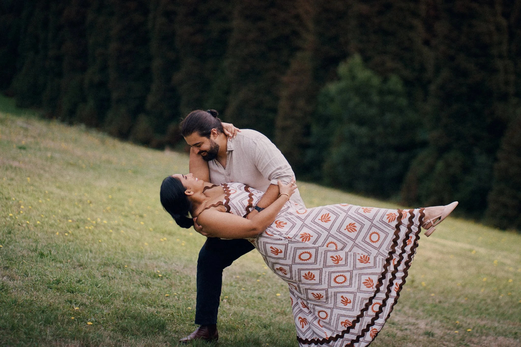 A man in a light-colored shirt and dark pants holding a woman in a patterned dress in a dip pose outdoors, surrounded by greenery.