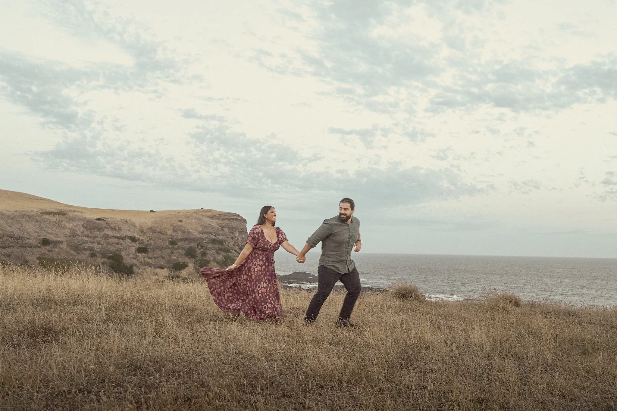 A couple holding hands and running through a grassy field near the ocean with a cloudy sky above.