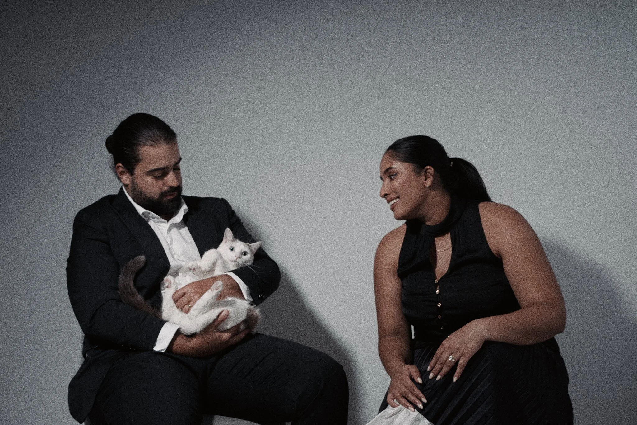 A man in a black suit holding a white and gray cat, and a woman in a black sleeveless top laughing and talking to him against a plain gray background.